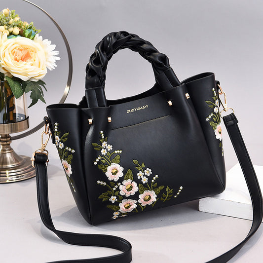 Black handbag with floral embroidery on a white surface with flowers in the background