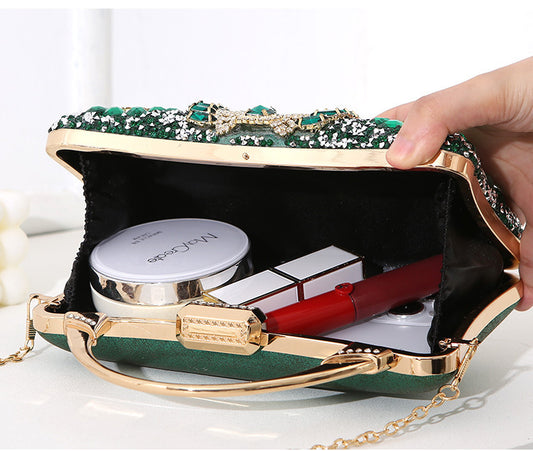 Green clutch bag with gold accents containing makeup items on a white surface.
