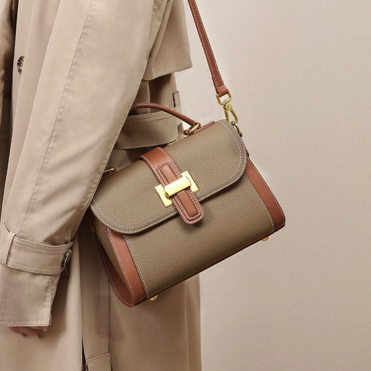 Brown handbag with gold clasp held by a person wearing a beige trench coat on a neutral background