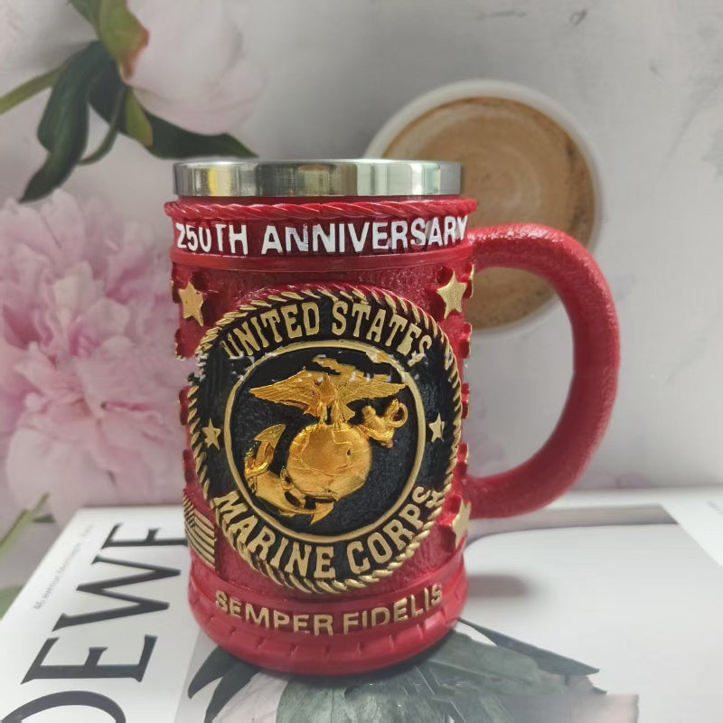 Red mug with United States Marine Corps logo and '250th Anniversary' text on a surface with flowers and a book.
