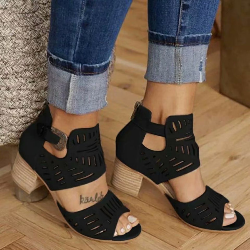 Black open-toe sandals with block heels worn with rolled-up jeans on a wooden floor.