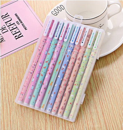 Set of colorful pens in a clear case on a wooden surface with a pink book and white dish in the background.