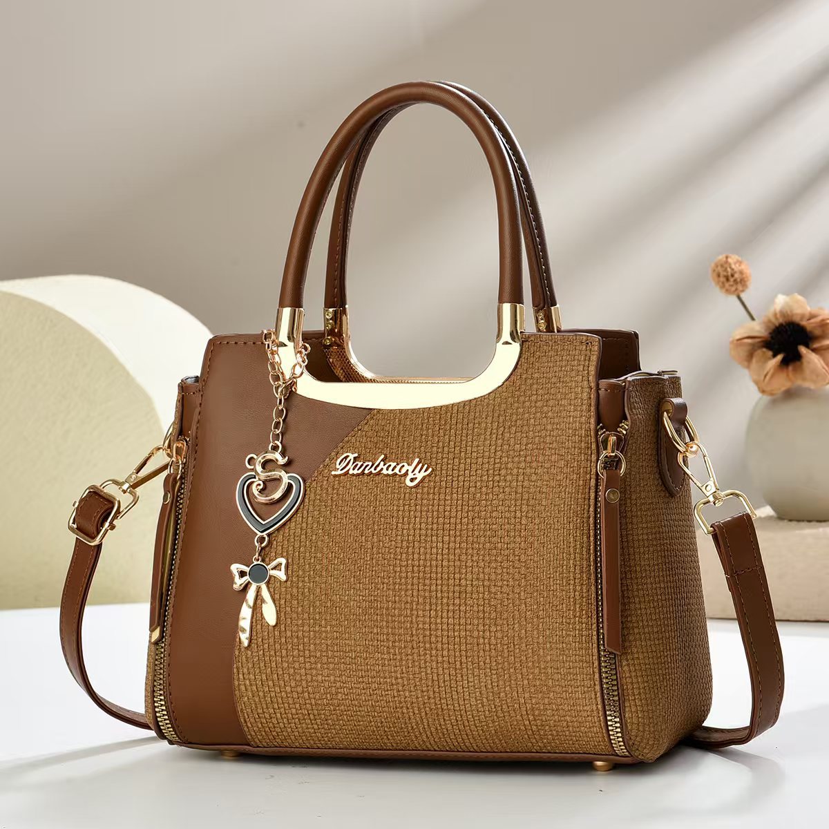 Small square crossbody bag, dark brown textured fabric, zipper, top handles, heart and bow charm.