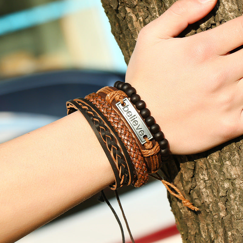 Close-up of a wrist wearing multiple leather bracelets with a 'believer' tag, against a blurred natural background.