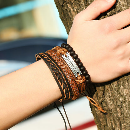 Close-up of a wrist wearing multiple leather bracelets with a 'believer' tag, against a blurred natural background.