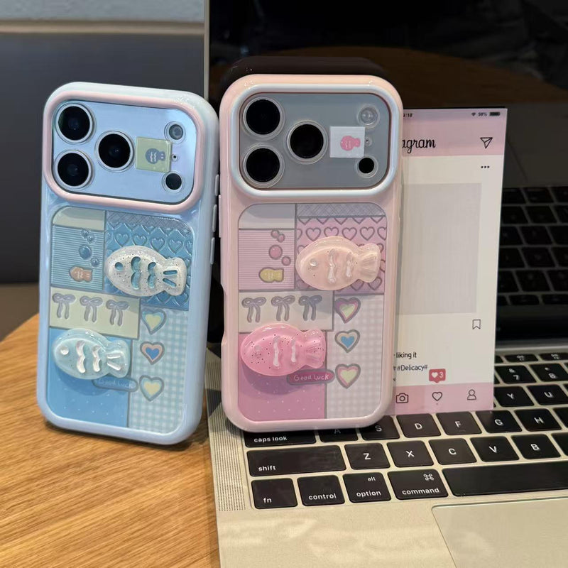 Two iPhone cases beside laptop: pale blue and pink with hearts and raised fish