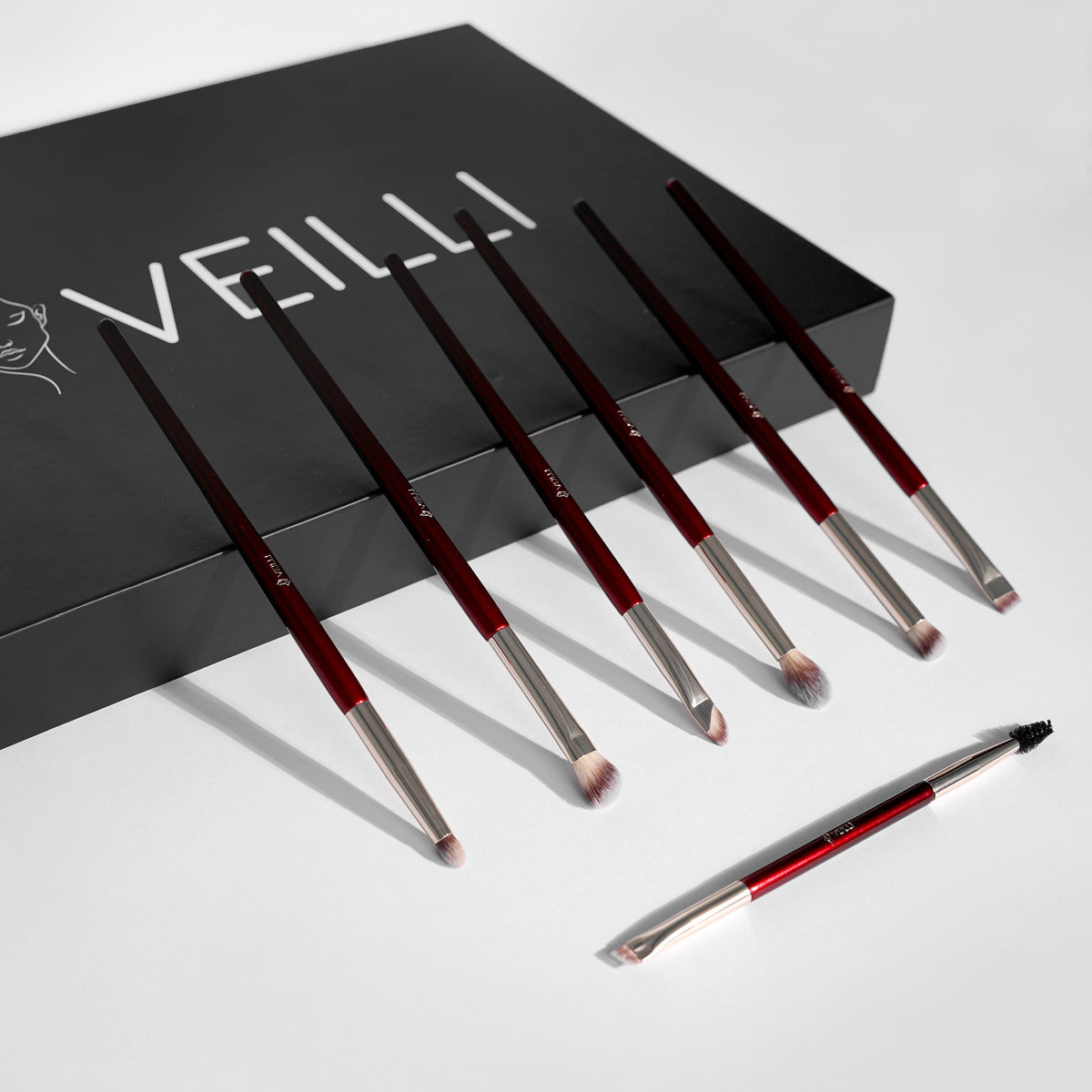 Slim burgundy makeup brushes with silver ferrules beside a black box