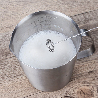 Electric milk frother whisking milk in a stainless steel measuring pitcher.