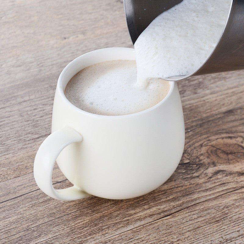 Electric milk frother shown pouring frothed milk from a stainless steel pitcher into a mug.