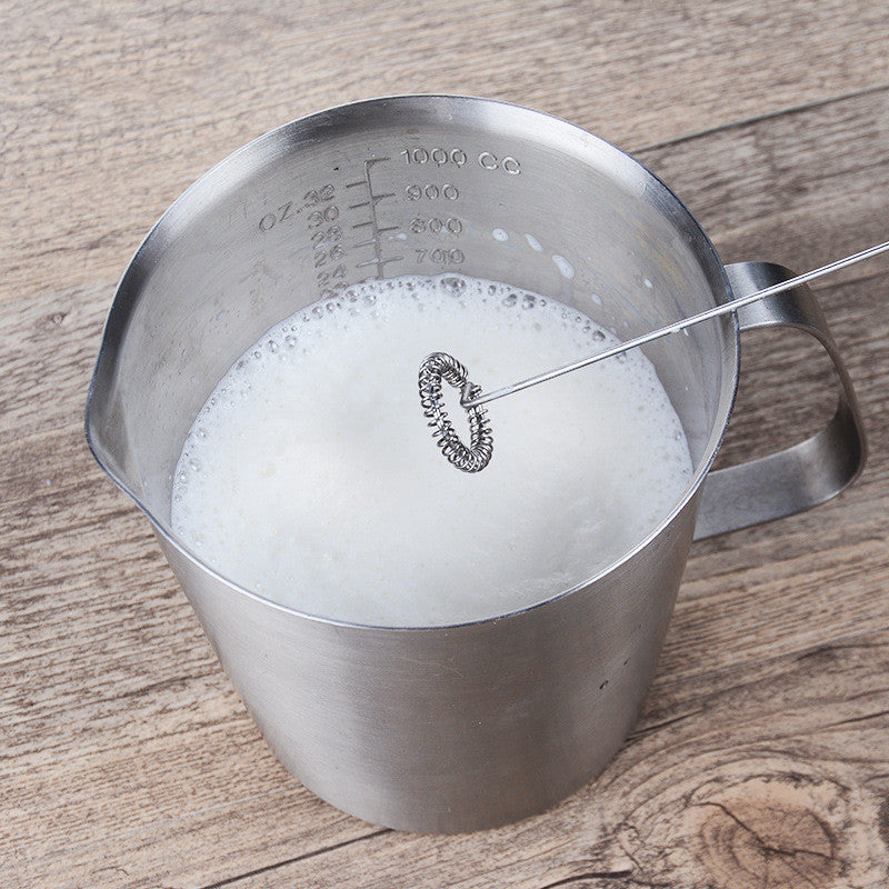 Electric milk frother whisking milk in a stainless steel measuring pitcher.