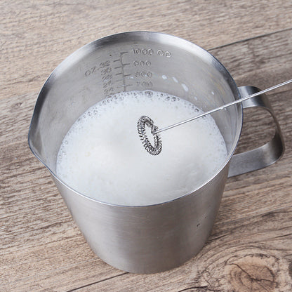 Electric milk frother whisking milk in a stainless steel measuring pitcher.